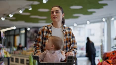 Close-up-a-confident-brunette-girl-in-a-plaid-shirt-carries-her-little-daughter-on-a-cart-while-shopping-and-inspecting-goods-in-a-supermarket