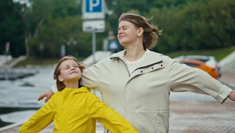 Happy-blonde-woman-together-with-her-teenage-daughter-girl-in-a-yellow-jacket-stand-against-the-wind-and-enjoy-the-wind-while-walking-in-the-park-after-the-rain