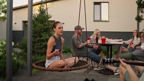 Happy-brunette-girl-sings-a-song-while-her-boyfriend-plays-the-guitar-during-a-party-during-the-day-in-the-backyard-of-a-country-house