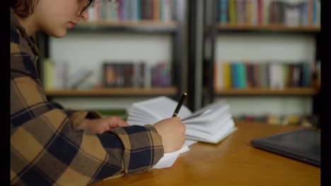 Close-up-of-a-girl-in-a-checkered-brown-shirt-takes-notes-at-a-table-in-the-university-library