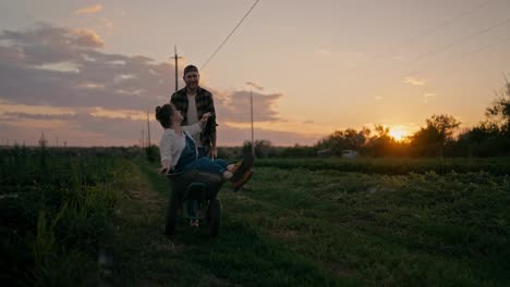 Ein-Glücklicher-Bauer-Schiebt-Seine-Freundin,-Einen-Bauern-In-Einem-Jeans-Overall,-In-Einer-Schubkarre-Am-Abend-Bei-Sonnenuntergang-Auf-Einem-Feld-Auf-Einem-Bauernhof