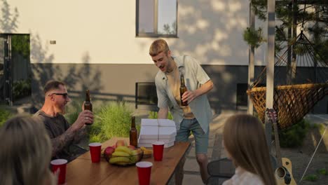 Happy-blond-guy-brings-a-package-of-pizza-and-bottles-of-drinks-to-the-table-during-a-shared-lunch-in-the-courtyard-of-a-country-house