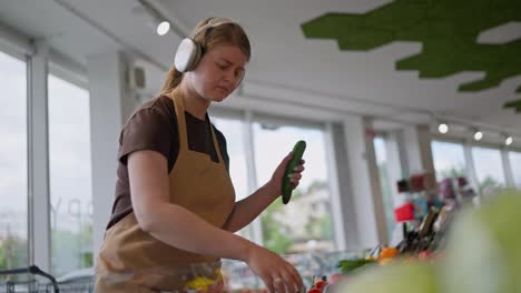 Confident-girl-in-a-brown-T-shirt-apron-and-headphones-arranges-vegetables-on-the-counter-and-listens-to-music-while-working-in-a-supermarket