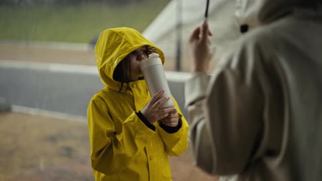 Por-Encima-Del-Hombro,-Una-Niña-Feliz-Con-Una-Chaqueta-Amarilla-Se-Comunica-Con-Su-Madre-Y-Bebe-Agua-De-Una-Botella-Blanca-En-El-Parque-Durante-Una-Caminata-Y-Una-Fuerte-Lluvia.