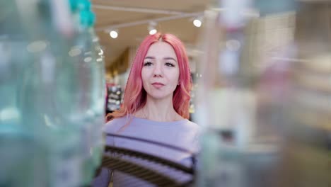 Close-up-a-confident-girl-with-pink-hair-in-a-purple-T-shirt-approaches-a-counter-with-bottles-of-water-and-chooses-the-right-one-for-herself-in-a-supermarket