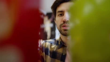 Close-up-a-confident-brunette-guy-with-stubble-in-a-checkered-shirt-approaches-the-counter-in-a-supermarket-and-chooses-the-red-product-he-needs