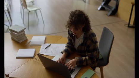 Top-view-of-a-concentrated-girl-with-curly-hair-sitting-at-a-table-and-doing-student-work-on-a-laptop-in-the-library