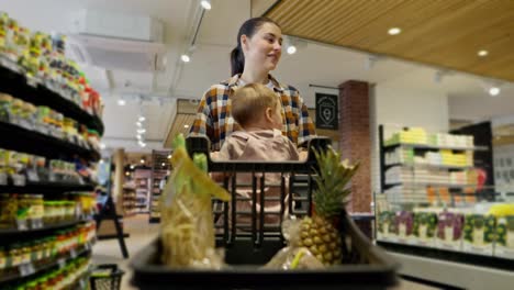 A-confident-brunette-girl-in-a-plaid-shirt-carries-a-cart-with-groceries-in-which-her-little-daughter-is-sitting-during-family-shopping-in-a-supermarket