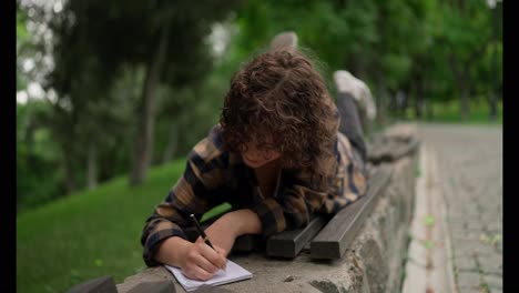 Happy-brunette-girl-with-curly-hair-makes-notes-in-her-notebook-while-lying-on-a-bench-in-the-park