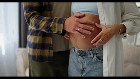 Close-up-of-a-happy-man-in-a-plaid-shirt-stroking-his-wife's-pregnant-belly-at-home-while-touching-her-hand