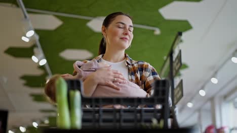 Bottom-view-of-a-happy-brunette-girl-with-a-baby-in-her-arms-looking-around-during-her-shopping-in-a-supermarket