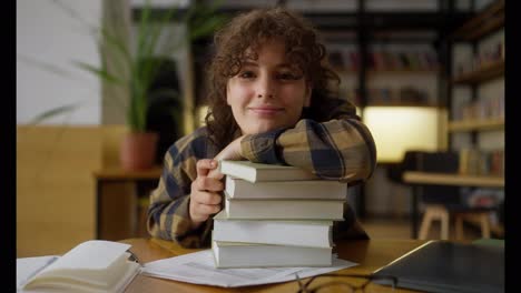 Retrato-De-Una-Estudiante-Feliz-Con-Cabello-Rizado-Que-Sonríe-Y-Se-Apoya-En-Una-Pila-De-Libros-Sobre-Una-Mesa-En-La-Biblioteca.