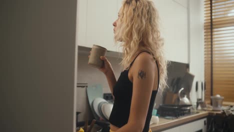 A-blonde-girl-in-a-black-top-drinks-tea-in-the-morning-and-looks-at-her-phone-in-the-morning-in-the-kitchen-near-the-refrigerator