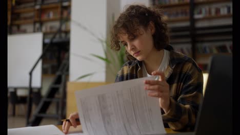 A-girl-student-with-piercings-with-curly-hair-in-a-checkered-shirt-sits-at-a-table-and-reads-protocols-for-work-in-the-university-library