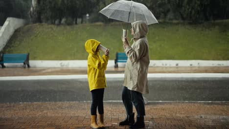 Happy-teenage-girl-in-a-yellow-jacket-drinks-water-and-communicates-with-her-mom-while-standing-under-an-umbrella-in-the-park-during-heavy-rain