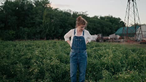 Confident-girl-Farmer-in-denim-overalls-with-red-curly-hair-inspecting-plants-in-a-field-on-a-farm