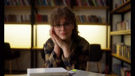 Portrait-of-a-happy-brunette-student-girl-in-glasses-and-a-plaid-shirt-who-sits-at-a-table-among-books-in-the-library