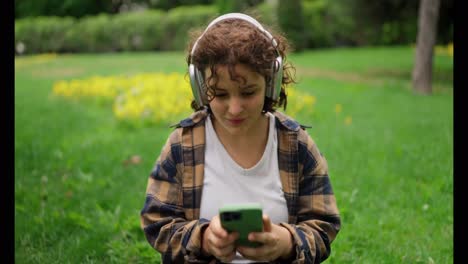 Close-up-of-a-happy-girl-with-curly-hair-in-white-wireless-headphones-resting-and-listening-to-music-in-the-park