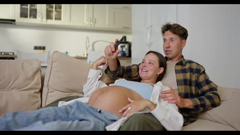 A-man-in-a-plaid-shirt-and-his-pregnant-wife-are-relaxing-on-the-sofa-and-laughing-while-watching-TV-at-home