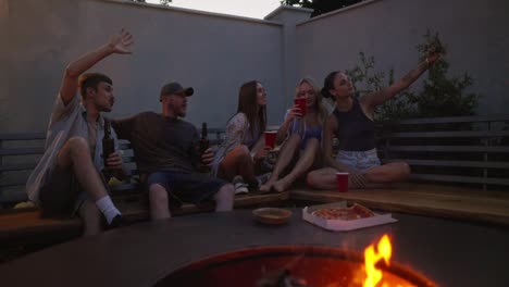 A-happy-and-cheerful-group-of-friends-takes-a-selfie-while-sitting-on-a-bench-near-a-barbecue-with-a-fire-during-their-party-in-the-evening-in-the-backyard-of-a-country-house