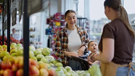 A-happy-brunette-girl-approaches-a-supermarket-worker-and-asks-about-the-vegetables-on-the-counter-during-her-shopping-with-her-daughter