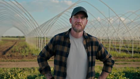 Portrait-of-a-happy-guy-farmer-with-a-beard-in-a-cap-who-poses-near-buildings-and-greenhouses-on-the-farm