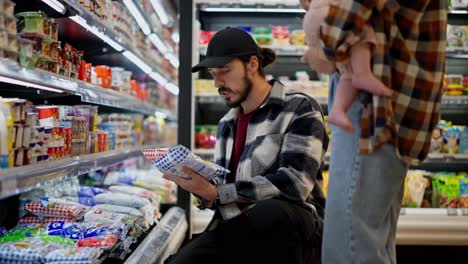 Brunette-guy-consults-with-his-wife-about-the-choice-of-dairy-products-during-family-shopping-in-the-supermarket.-A-brunette-girl-together-with-her-husband-and-infant-choose-goods-in-a-supermarket