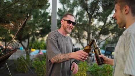 Happy-guy-in-sunglasses-with-beard-clinks-brown-bottles-with-his-friend-while-his-girlfriend-approaches-and-joins-conversation-in-backyard-while-relaxing