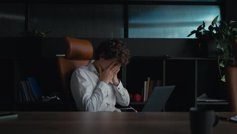 Side-view-of-a-tired-guy-with-curly-hair-in-a-white-shirt-covers-his-face-with-his-hands-and-shakes-his-head-during-a-hard-day-at-work-in-the-office