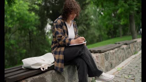 A-girl-student-with-curly-hair-in-a-brown-shirt-sits-on-a-bench-and-makes-notes-in-a-notebook-in-the-park