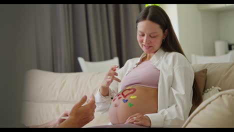 Side-view-of-a-happy-brunette-pregnant-woman-drawing-hearts-on-her-belly-using-bright-colors-together-with-her-husband-while-expecting-a-baby-at-home