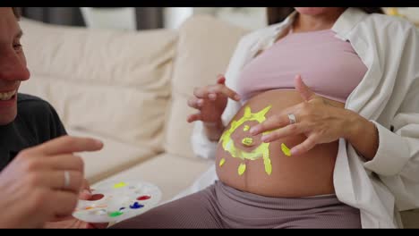 Close-up-of-a-happy-pregnant-woman-drawing-a-sun-on-her-belly-while-having-fun-with-her-husband-at-home-on-the-sofa