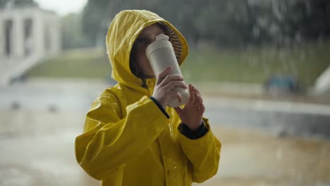 A-blonde-teenage-girl-in-a-yellow-jacket-drinks-water-from-a-white-plastic-bottle-and-looks-at-the-heavy-rain-in-the-park