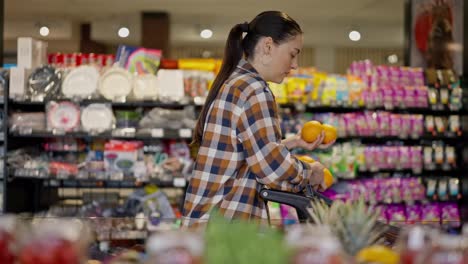 A-brunette-girl-in-a-plaid-shirt-puts-fruits-in-a-cart-during-her-shopping-in-a-supermarket.-Confident-brunette-woman-chooses-oranges-during-her-shopping