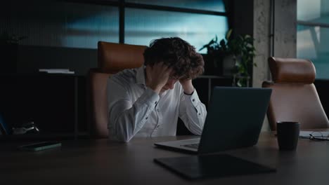 Sad-and-tired-office-worker-guy-with-curly-hair-puts-his-head-on-the-laptop-keyboard-and-shakes-it-to-the-side-during-a-difficult-task-in-the-office