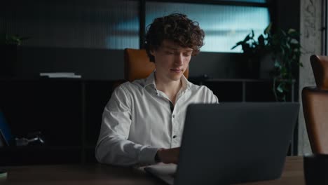 Confident-young-guy-with-curly-hair-in-a-white-shirt-typing-on-a-gray-laptop-while-working-in-the-office