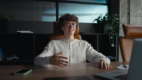 Un-Chico-Feliz-Con-Cabello-Rizado-Y-Gafas-Con-Una-Camisa-Blanca-Se-Sienta-A-La-Mesa-Y-Se-Comunica-Por-Videoconferencia-Usando-Una-Computadora-Portátil-En-La-Oficina-Durante-La-Jornada-Laboral.