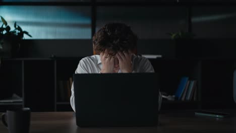 Un-Chico-De-Pelo-Rizado-Con-Una-Camisa-Blanca-Se-Cubre-La-Cara-Con-Las-Manos-Y-Mueve-La-Cabeza-Hacia-Los-Lados-Durante-Una-Tarea-Difícil-En-El-Trabajo-En-La-Oficina.