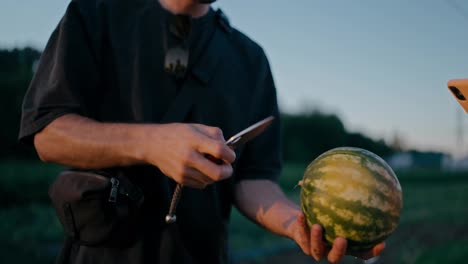 Nahaufnahme-Eines-Mannes-In-Einem-Schwarzen-T-Shirt,-Der-Auf-Einem-Feld-Auf-Einer-Farm-Eine-Kleine-Wassermelone-Aufschneidet-Und-Die-Hälfte-Seinem-Freund-Gibt