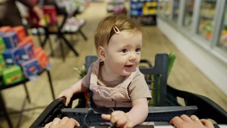 Portrait-of-a-small-infant-child-who-sits-in-a-cart-during-family-shopping-in-a-supermarket.-A-little-girl-looks-at-everything-around-her-while-shopping-with-her-parents-in-the-supermarket