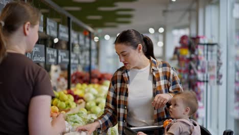 Over-the-shoulder-a-happy-brunette-girl-with-her-little-child-asks-a-supermarket-worker-about-the-products-on-the-vegetable-counter