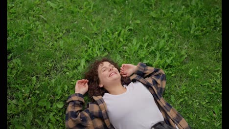 Happy-brunette-girl-with-curly-hair-lies-with-her-back-on-the-green-lawn-in-the-park