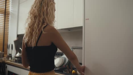 Rear-view-of-a-blonde-girl-with-curly-hair-drinking-coffee-and-looking-into-the-refrigerator-with-a-phone-in-her-hands-in-the-morning