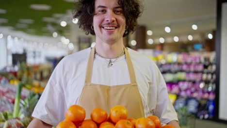 Porträt-Eines-Glücklichen-Brünetten-Kerls-Mit-Lockigem-Haar,-Der-In-Seinen-Händen-Einen-Korb-Mit-Vielen-Orangefarbenen-Zitrusfrüchten-In-Einem-Supermarkt-Hält