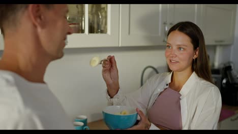 Happy-brunette-woman-feeding-her-husband-with-a-fork-fruit-salad-in-the-kitchen-in-the-morning