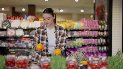 A-brunette-girl-in-a-checkered-shirt-chooses-one-of-two-oranges-in-the-supermarket-department-while-shopping