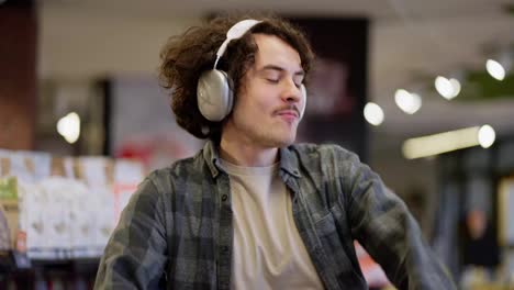 Close-up-of-a-happy-brunette-guy-with-curly-hair-wearing-white-headphones-dancing-and-listening-to-music-during-his-shopping-in-the-supermarket