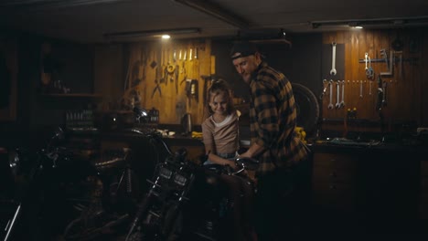 Happy-guy-mechanic-in-a-checkered-shirt-shows-his-workshop-and-motorcycles-to-his-little-daughter