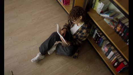 Top-view-of-a-girl-student-with-curly-hair-in-a-plaid-shirt-reading-a-book-near-shelves-with-books-in-the-library