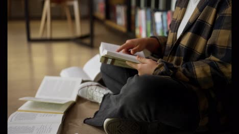 Nahaufnahme-Einer-Studentin-In-Einem-Karierten-Hemd,-Die-Ein-Buch-Liest,-Während-Sie-Auf-Dem-Boden-Zwischen-Büchern-In-Der-Bibliothek-Sitzt